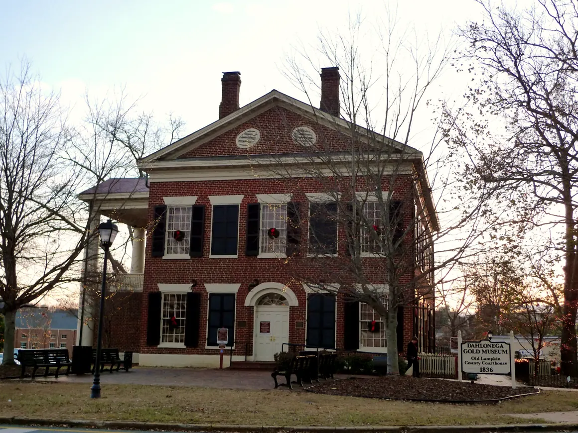 Gable end of the red-brick Dahlonega Gold Museum with wreaths in its windows, bare trees strung with white lights, and its 1836 sign.
