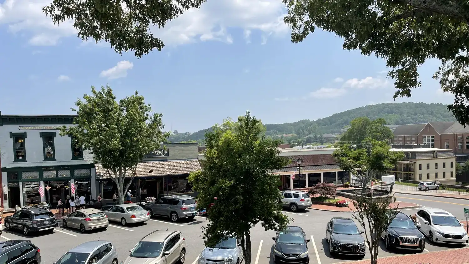 Elevated view of Dahlonega's public square in summer: storefronts, a parking lot full of cars, and forested hills behind under blue sky.