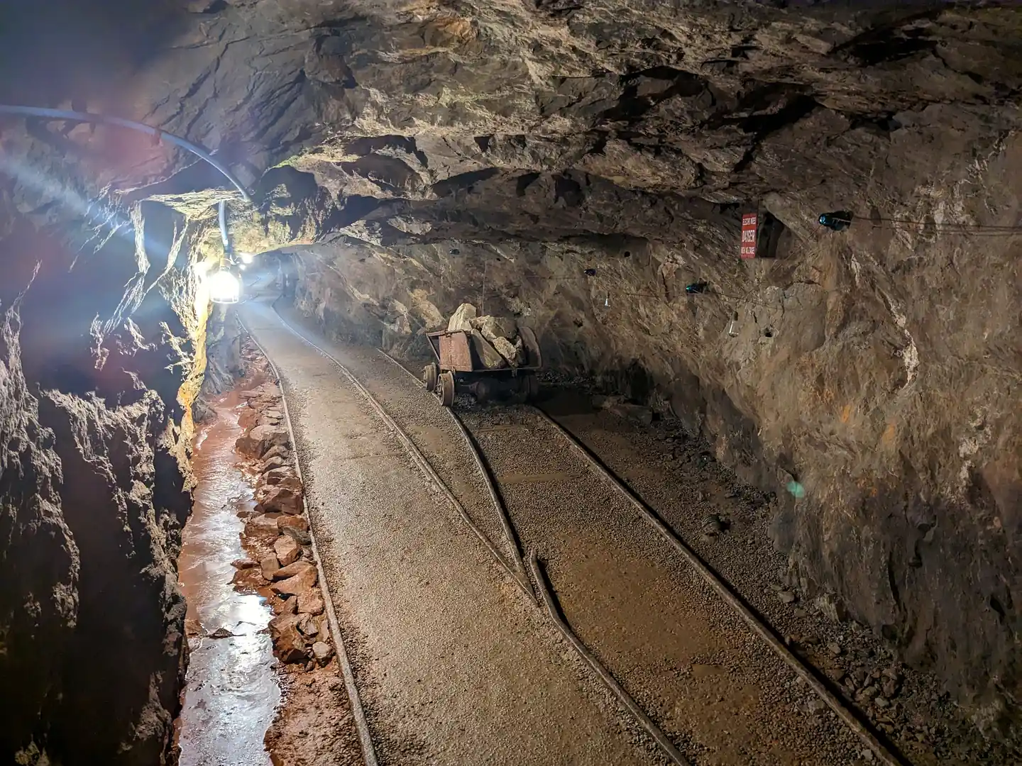 Underground tunnel at Consolidated Gold Mine with rails on a gravel floor, a rusty ore cart of rocks and a hanging work lamp.