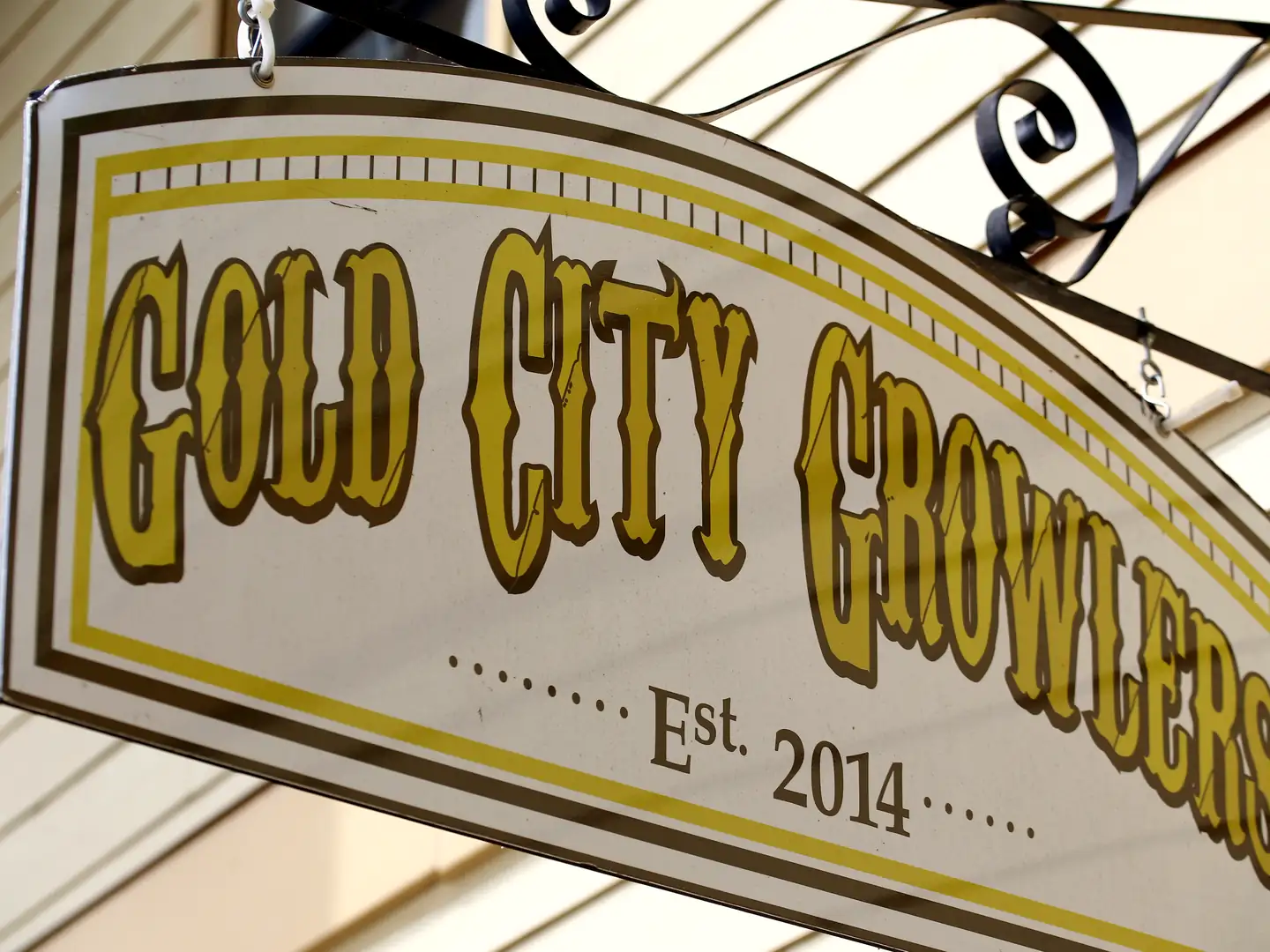Hanging sign reading 'Gold City Growlers, Est. 2014' on a black scrolled iron bracket against cream siding.