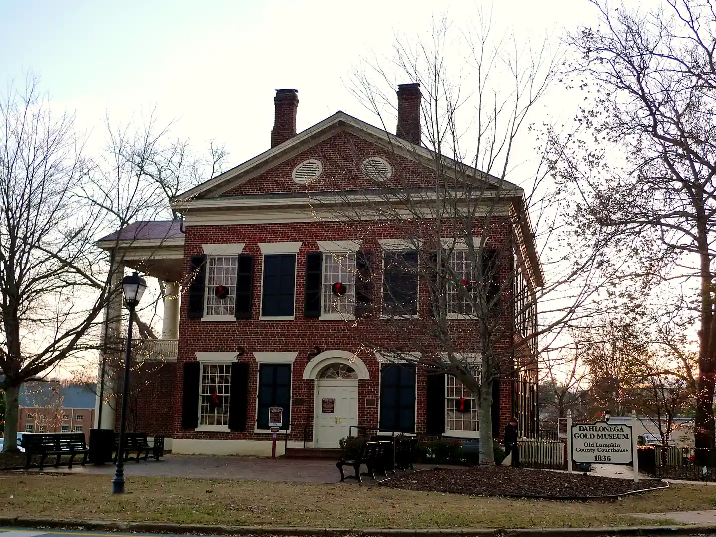 Gable end of the red-brick Dahlonega Gold Museum with wreaths in its windows, bare trees strung with white lights, and its 1836 sign.