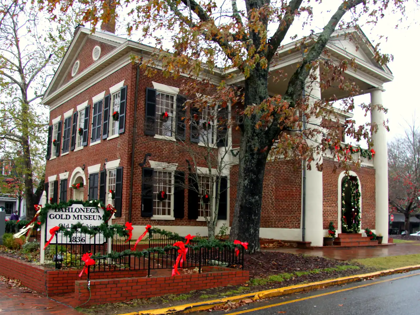 Red-brick Dahlonega Gold Museum with Christmas wreaths in the windows, garland on the fence and doorway, and its 1836 sign out front.