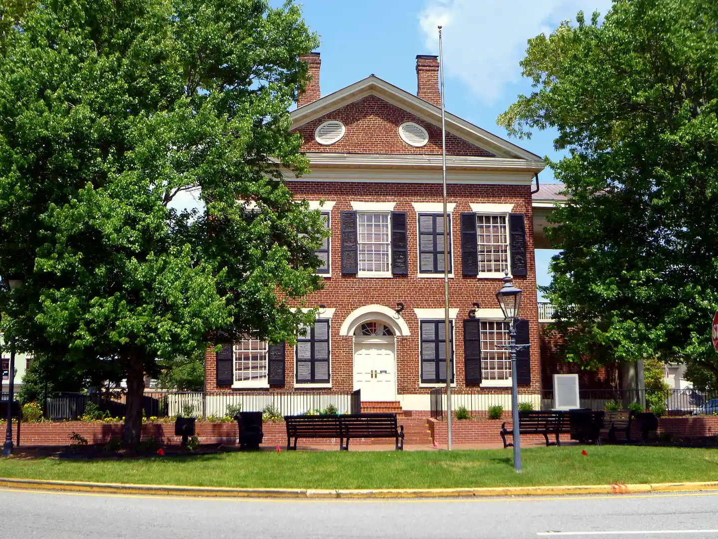Gable end of the red-brick Dahlonega Gold Museum framed by two leafy trees, with benches and a mown lawn in front.