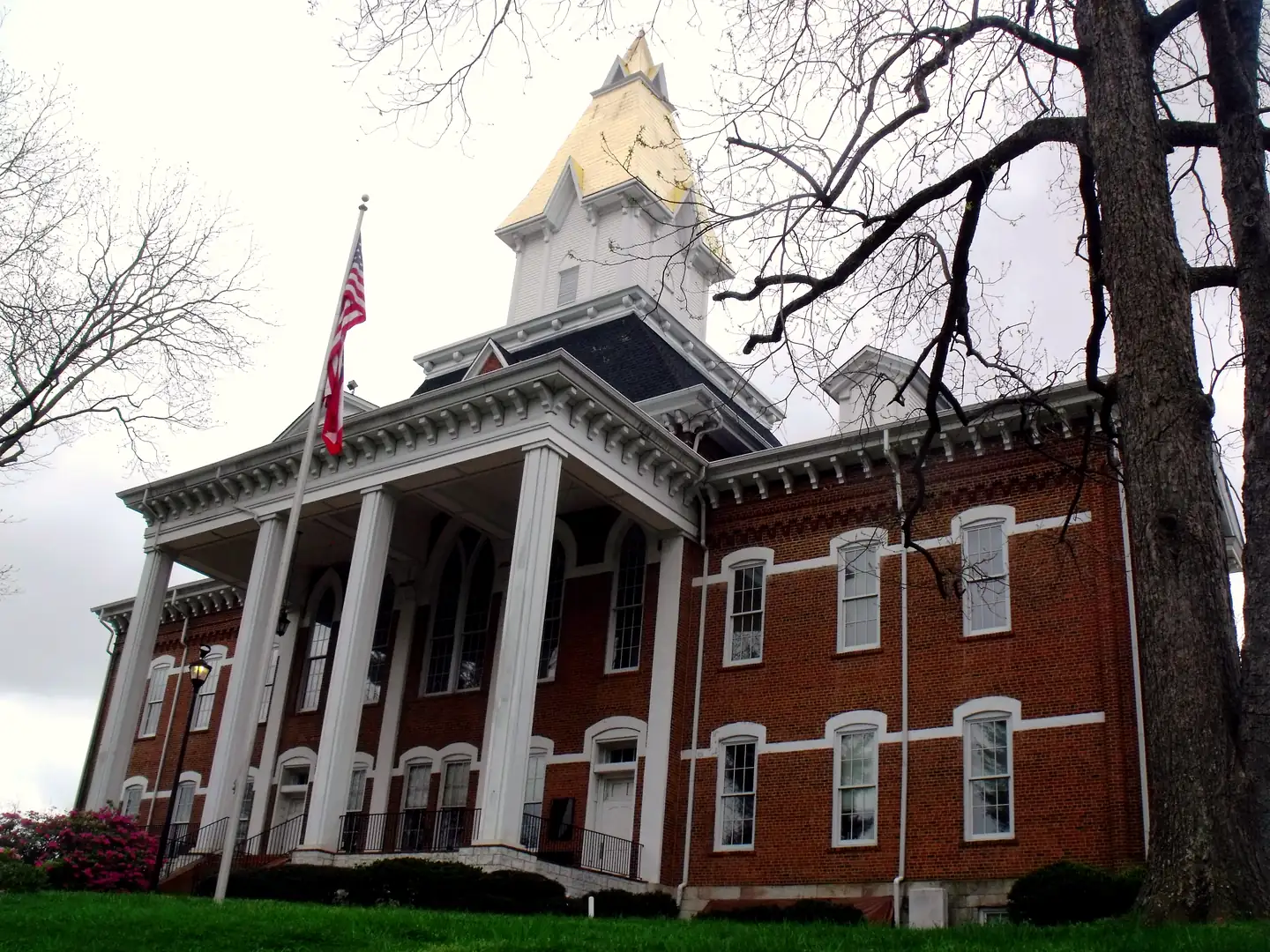 Low-angle view of Price Memorial Hall at UNG: red brick, four white columns, and a white belfry with a gold steeple, under an overcast sky.