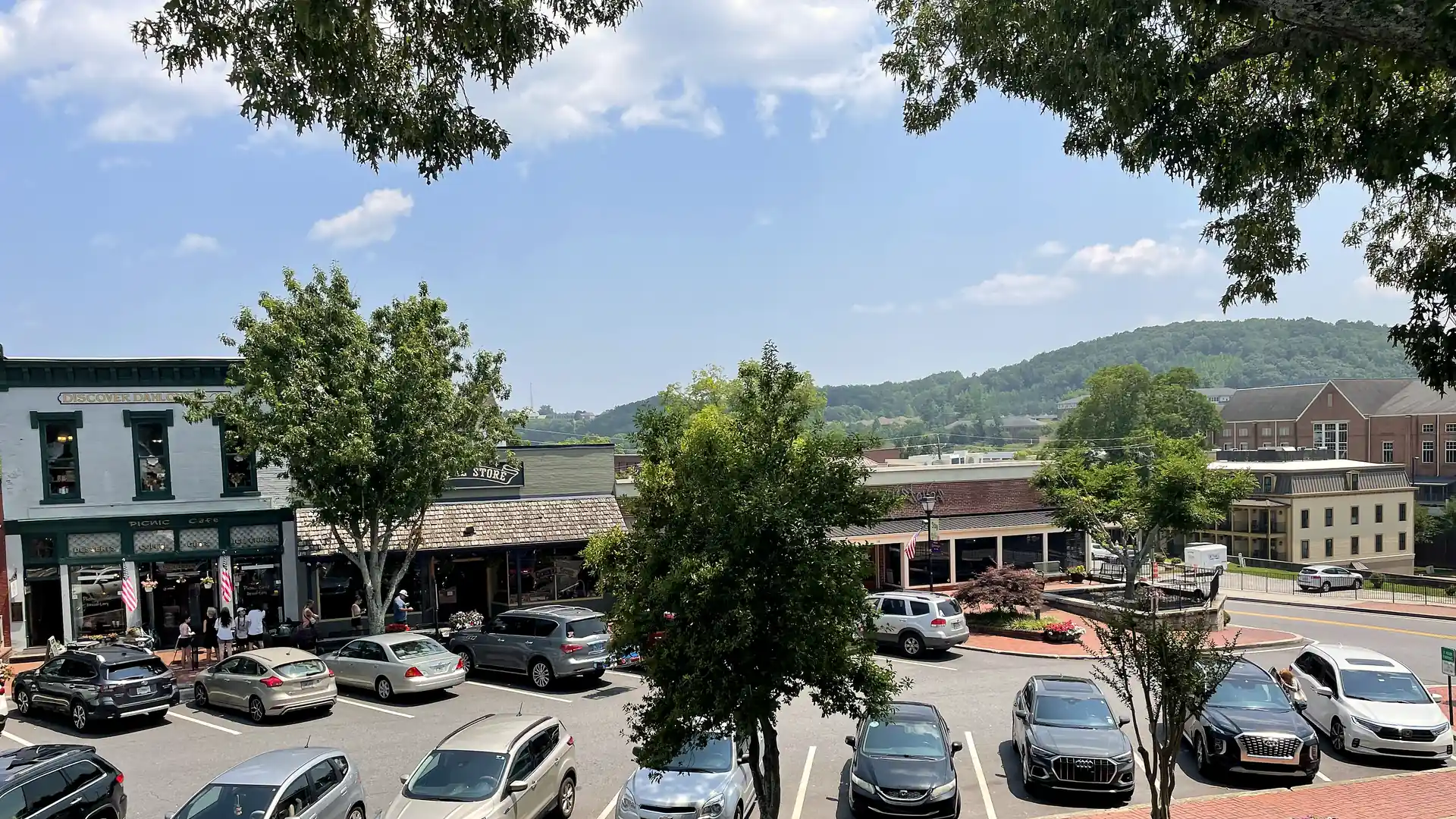 Elevated view of Dahlonega's public square in summer: storefronts, a parking lot full of cars, and forested hills behind under blue sky.
