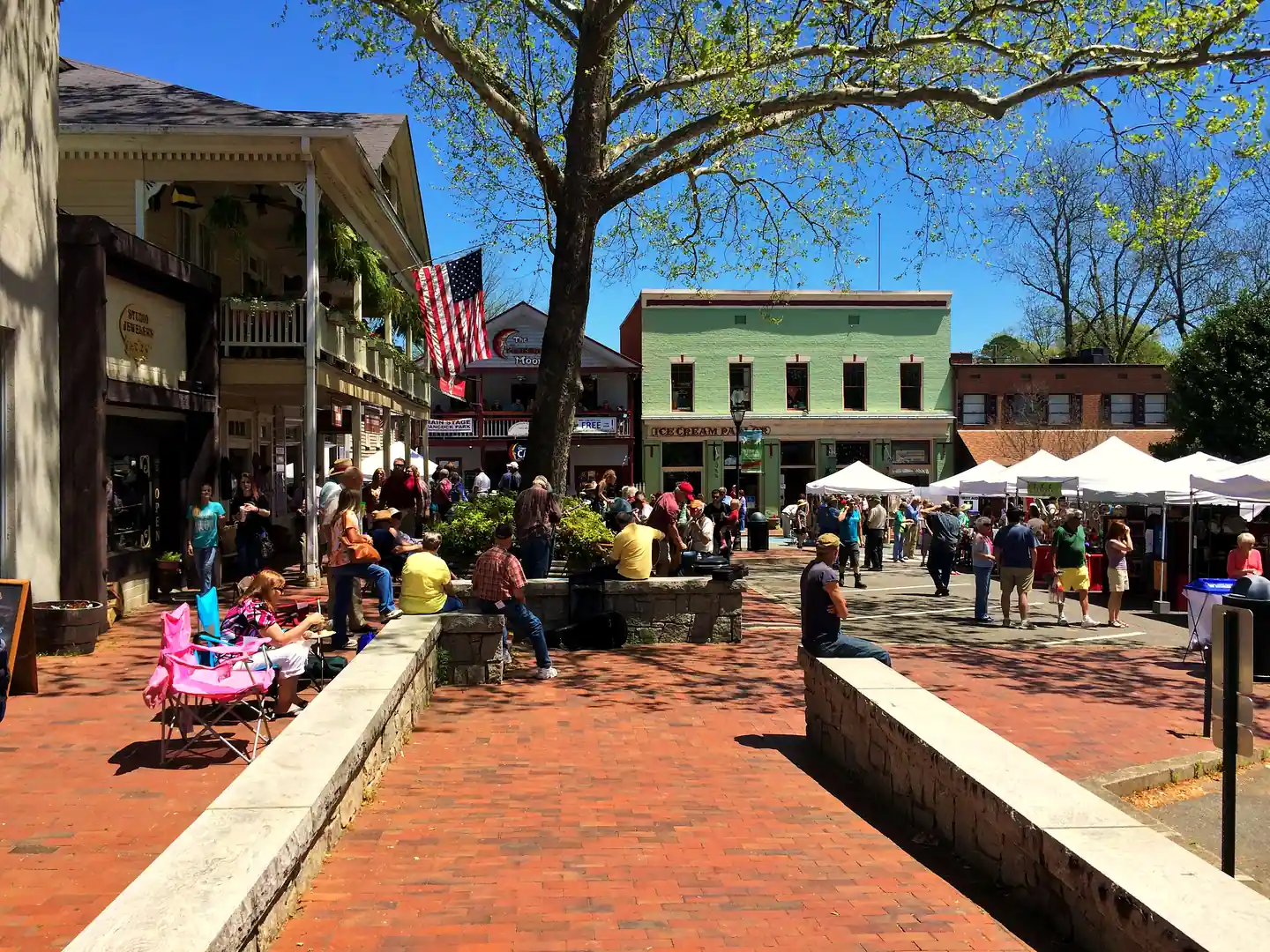 Crowded brick plaza on Dahlonega's public square on a sunny day, with an ice cream parlor, a Crimson Moon sign, white vendor tents and a large sycamore.